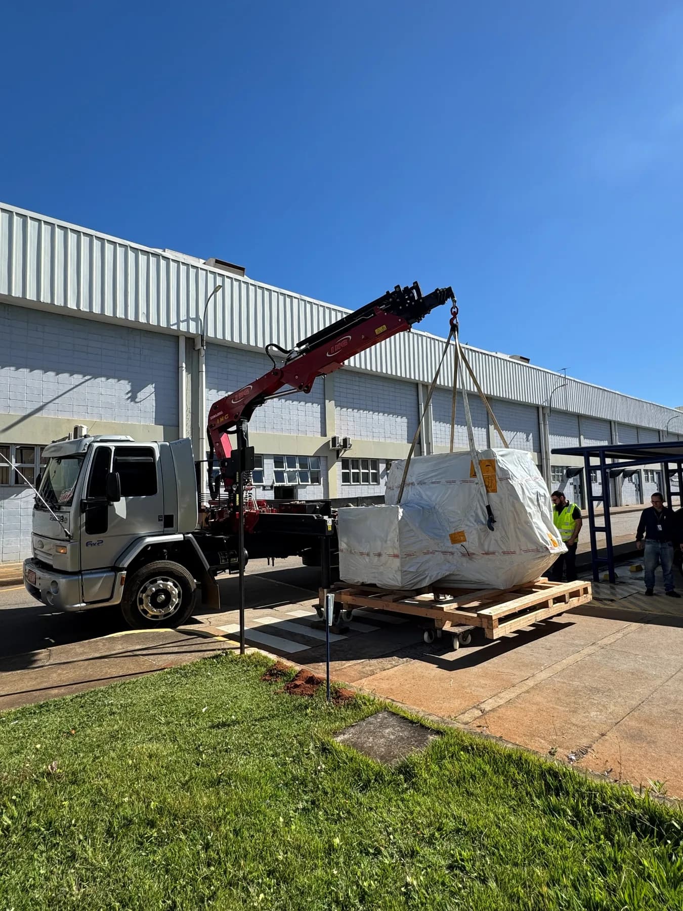 Munck içando por cintas um equipamento embalado sobre pallet em frente a um galpão industrial, acompanhado por dois funcionários