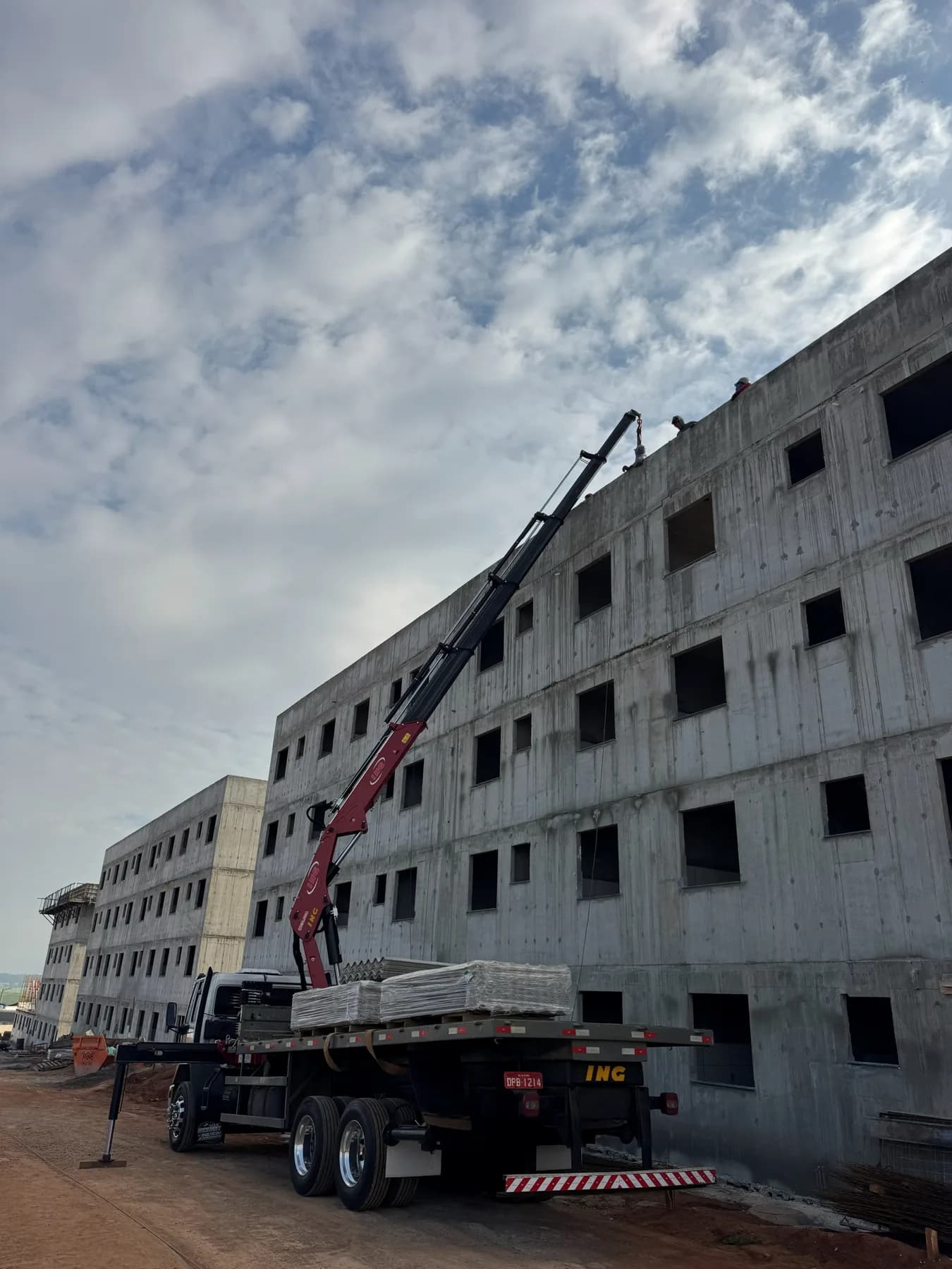 Lança do munck estendida até o quarto pavimento de um prédio de concreto em construção, entregando um fardo de material a trabalhadores na laje