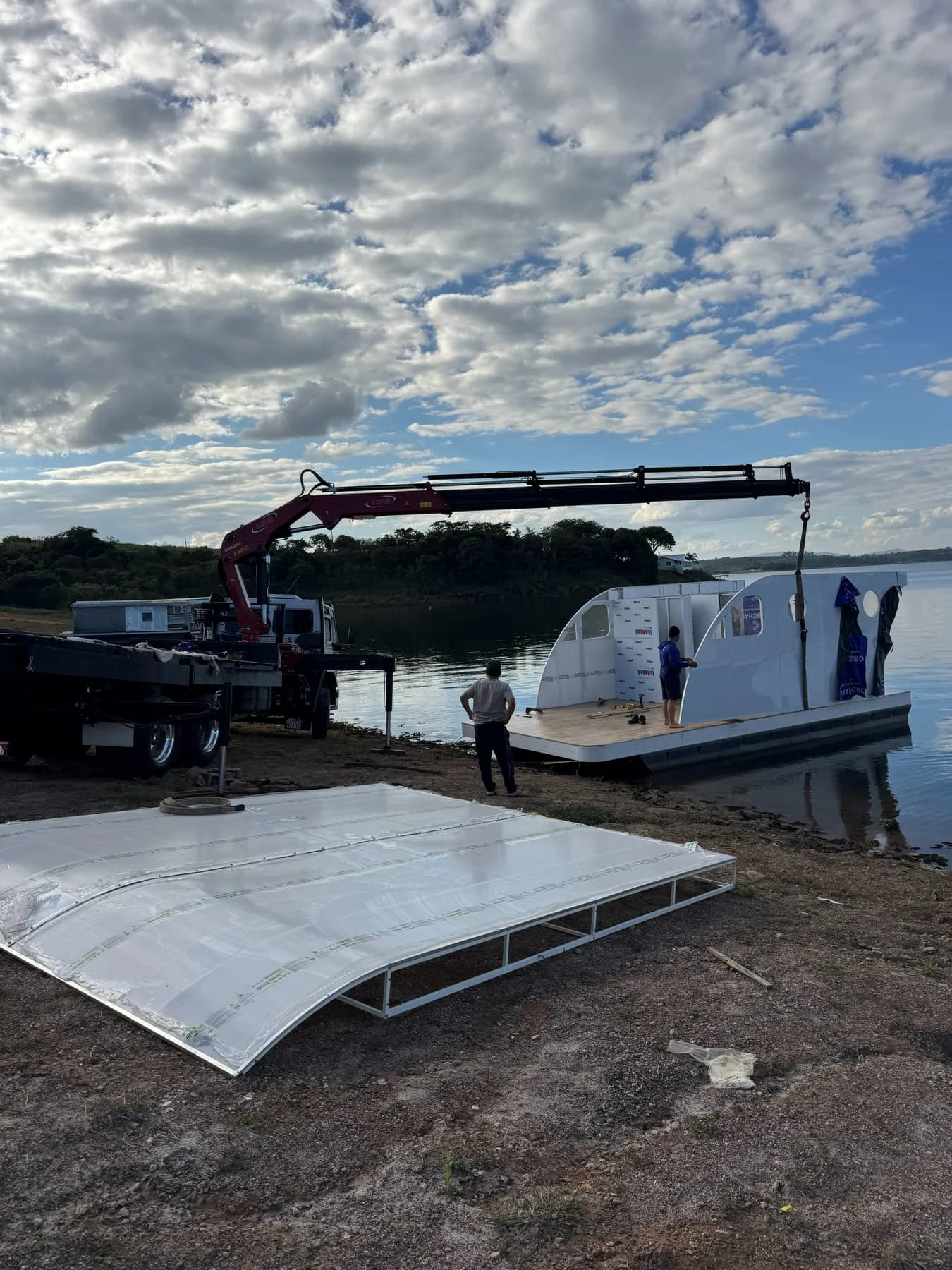 Munck montando a estrutura metálica de um flutuante na margem de um lago, com a peça de cobertura ainda no chão