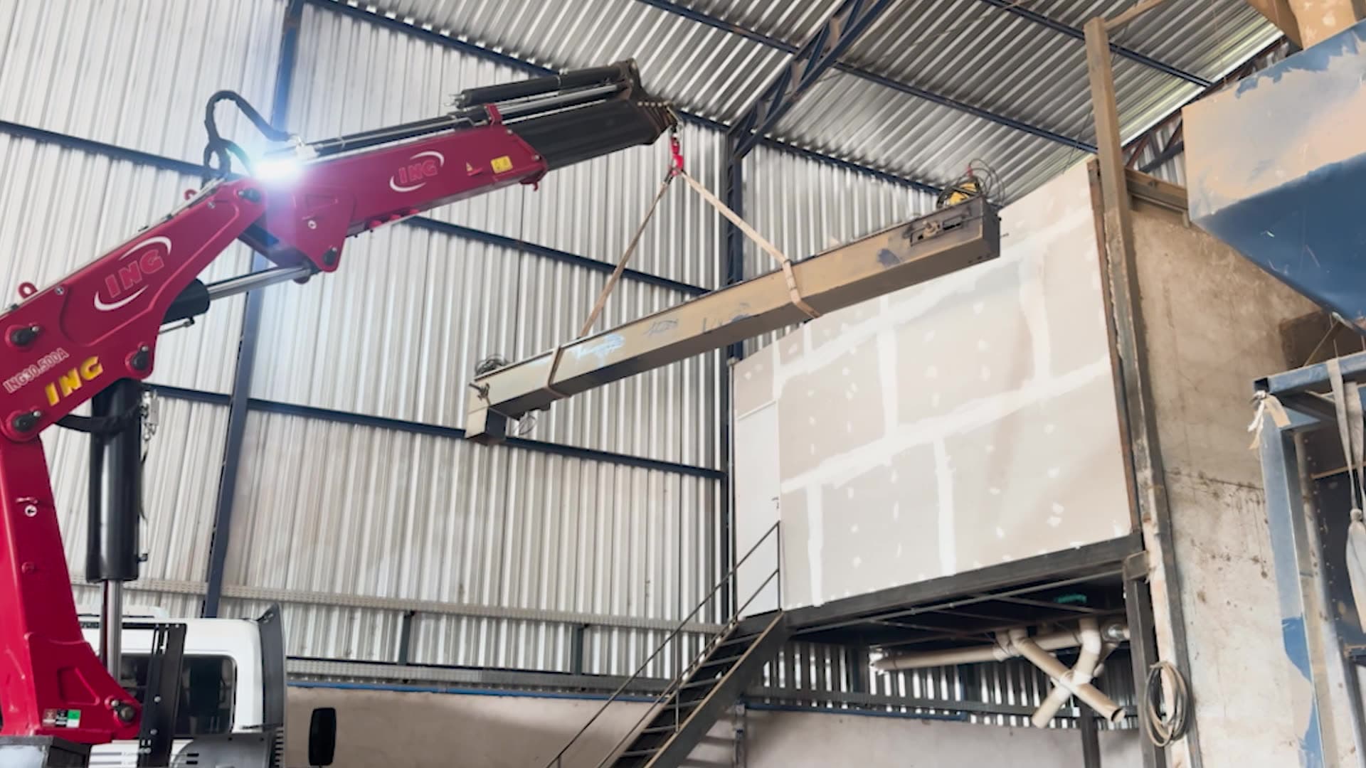 Lança vermelha do caminhão munck ING estendida dentro de um galpão industrial, sustentando por cintas uma viga metálica longa na horizontal